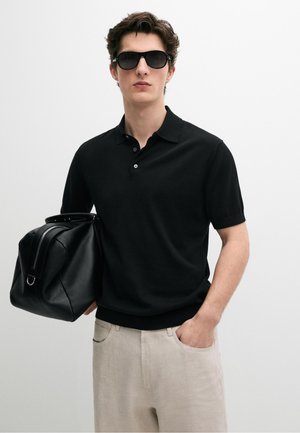 Jonge man met een zwart poloshirt en zonnebril, beige broek, houdt een zwarte leren tas vast tegen een effen achtergrond.