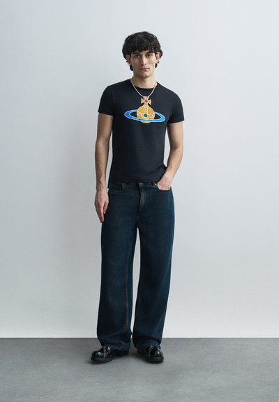 Camiseta negra con un gráfico colorido de una nave espacial orbitando un planeta, combinada con jeans de mezclilla oscura de pierna ancha y zapatos negros.