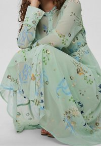 Robe longue vert menthe en tissu transparent, ornée de broderies florales de différentes couleurs et de détails dorés, avec un design à boutons.