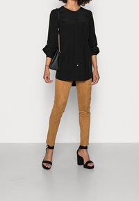 Blusa nera con bottoni e maniche lunghe a sbuffo, abbinata a pantaloni slim in suede marrone e sandali neri con tacco alto.