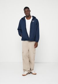 Tânăr îmbrăcat cu o jachetă Reebok bleumarin cu fermoar, tricou alb, pantaloni cargo bej și adidași albi, stând în fața unui fundal alb simplu.