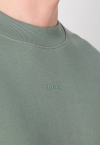 Gros plan d'une personne portant un t-shirt côtelé vert à col rond avec le mot HUGO embossé sur la poitrine.