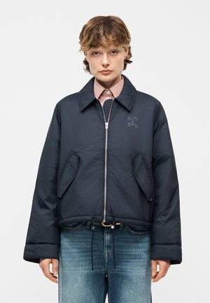 Osoba s krátkymi vlasmi oblečená v námorníckom modrom zipsovým bundom s prednými vreckami a nápisom na hrudi, modrými džínsami a pruhovanou košeľou s golierom.