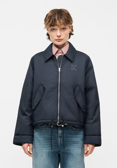 Person mit kurzen Haaren, die eine marineblaue Jacke mit Reißverschluss und Fronttaschen sowie Text auf der Brust trägt, blaue Jeans und ein gestreiftes Hemd mit Kragen.