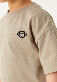 Beiges Baumwoll-T-Shirt mit einem schwarzen gestickten Affenlogo auf der Brust, Rundhalsausschnitt und kurzen Ärmeln mit glatter Textur.