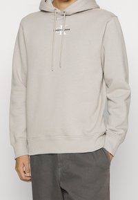 Beige mikina s kapucí vyrobená z měkkého materiálu. Má přední logo, stahovací šňůrku a žebrované manžety a lem pro zvýraznění detailů a správné padnutí.