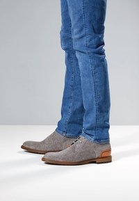 Grijs gemêleerde stoffen schoenen met bruine lederen accenten, gecombineerd met blauwe spijkerbroeken. De schoenen hebben een ronde neus en veters.