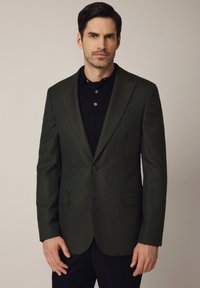 Ciemnozielony wełniany żakiet o dopasowanym kroju, z pojedynczym zapięciem na guzik, klapami w kształcie litery notch oraz dwiema przednimi kieszeniami. Noszony na czarnej koszulce polo.