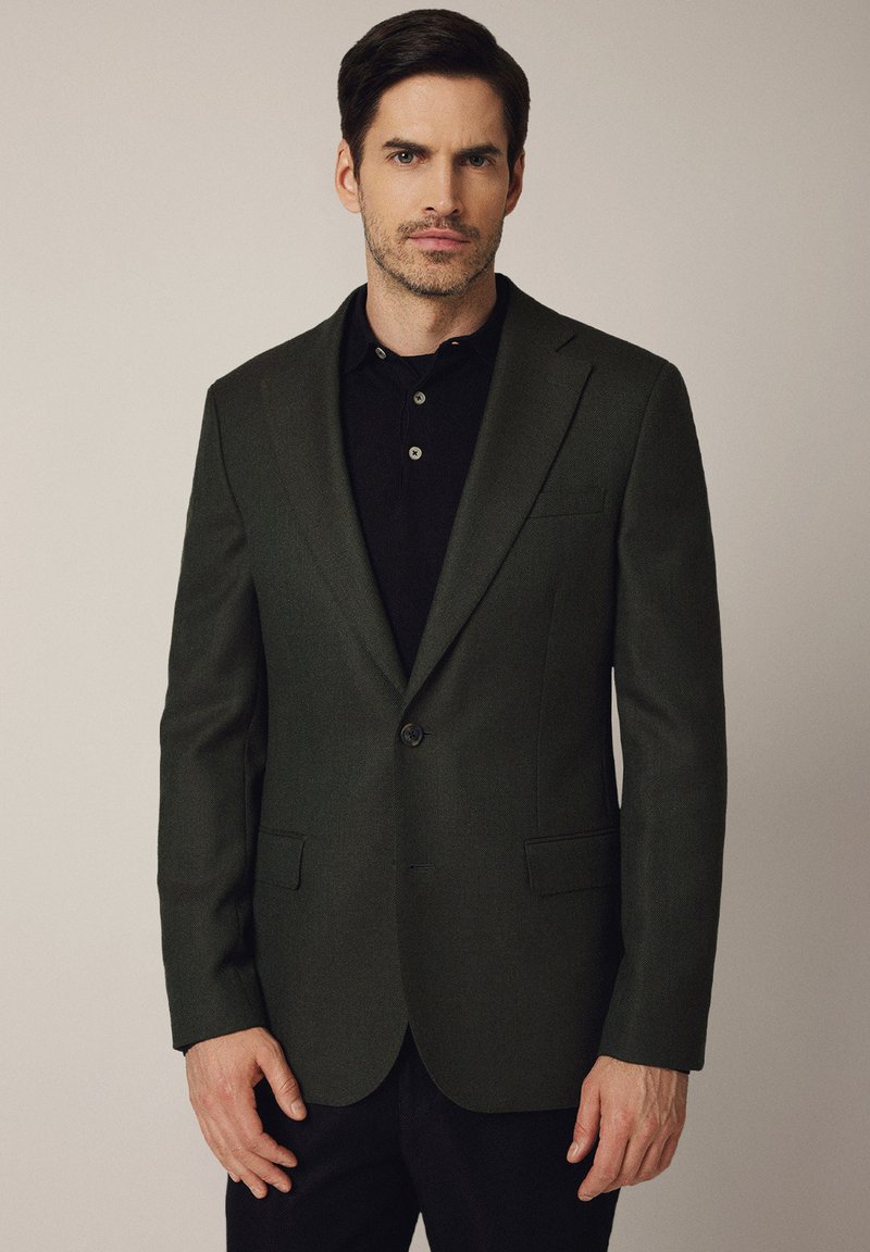 Ciemnozielony wełniany żakiet o dopasowanym kroju, z pojedynczym zapięciem na guzik, klapami w kształcie litery notch oraz dwiema przednimi kieszeniami. Noszony na czarnej koszulce polo.