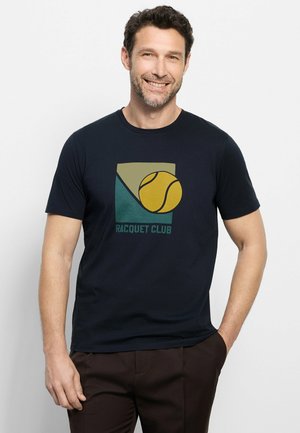 Lachende man die een zwart "Racquet Club" T-shirt draagt met een gele tennisbal en geometrische vormen, staand met één hand in de zak.