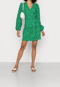 Vestido verde envolvente con estampado floral blanco, mangas abullonadas largas y un lazo en la cintura. Combinado con sandalias deslizantes color crema. Textura suave.