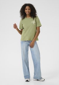 T-shirt en coton vert clair avec des manches courtes et un col rond, arborant un texte graphique blanc. Associé à un jean large bleu clair et des baskets.