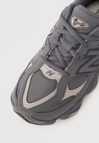 Gray athletic shoe with mesh and suede materials, featuring a textured surface, lacing system, and a reflective logo detail.