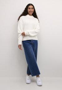 Maglione bianco a coste con collo alto, abbinato a jeans larghi blu scuro e sneakers bianche, in piedi contro uno sfondo chiaro.
