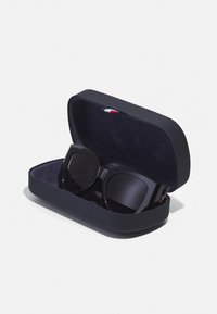 Ochelari de soare negri, supradimensionați, într-un etui rigid de culoare albastru marin. Interiorul etuiului are o textură moale, iar exteriorul are un mic steguleț colorat.