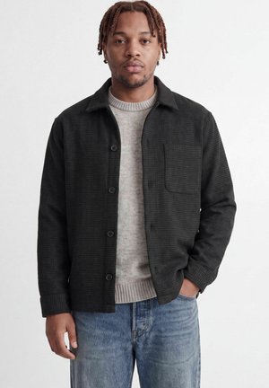 Mand med fletninger iført en sort skjorte med knapper over en lys sweater og blå jeans, stående mod en ensartet lys baggrund.