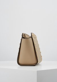 Beżowa torebka w kształcie geometrycznym z złotymi ćwiekami w kształcie piramidy po bokach oraz zamknięciem na zamek u góry. Gładki, teksturowany materiał.