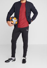 Idrottare i en marinblå jacka, vinröd skjorta, svarta byxor och svarta fotbollsskor, håller en grå-orange fotboll.