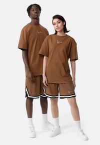 Brown oversized t-shirts and matching shorts with black accents. Both wear white sneakers and crew-length socks. Simple logo detail on shirts.