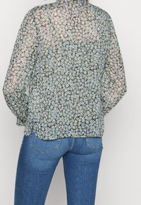 Blusa negra transparente con un estampado floral en azul y verde, con mangas largas, un cuello alto y un dobladillo curvado.