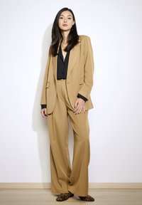 Beige tailor-made pak met wijde pijpen en een bijpassende blazer. Zwarte blouse eronder; schoenen met dierenprint. Glad materiaal en gestructureerd ontwerp.