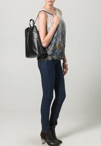 Sac à dos en cuir noir avec un design structuré, porté sur l'épaule. La personne porte un haut multicolore et un jean foncé ajusté avec des bottines.