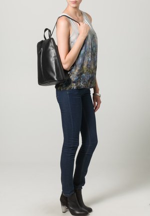 Sac à dos en cuir noir avec un design structuré, porté sur l'épaule. La personne porte un haut multicolore et un jean foncé ajusté avec des bottines.