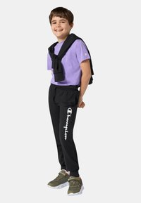 T-shirt viola a maniche corte, pantaloni da ginnastica neri con il logo "Champion", maglione nero annodato attorno alle spalle e scarpe sportive verdi.