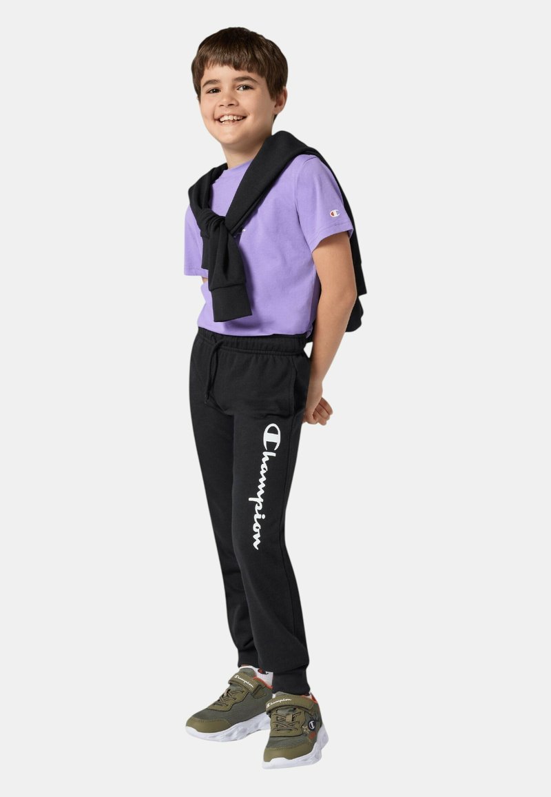 T-shirt viola a maniche corte, pantaloni da ginnastica neri con il logo "Champion", maglione nero annodato attorno alle spalle e scarpe sportive verdi.