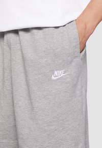 Osoba v světle šedých šortkách Nike s rukou v kapse, s logem Nike na levé stehně na bílém pozadí.