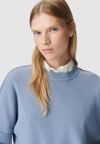 Pull en tricot bleu clair avec une texture côtelée, un col rond et une coupe décontractée. Porté sur une chemise blanche à col, avec des coutures minimales.