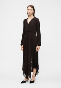 Γυναίκα που φοράει μαύρο πλισέ μεσάτο φόρεμα με μακριά μανίκια, V-λαιμόκοψη και ασύμμετρο τελείωμα, συνδυασμένο με μαύρα μυτερά ψηλοτάκουνα παπούτσια.