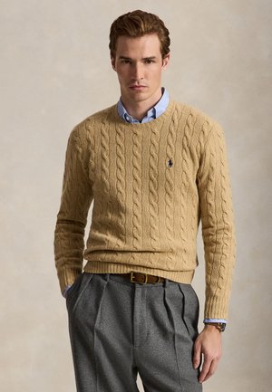 Homme portant un pull beige en maille torsadée sur une chemise bleue boutonnée, un pantalon gris plissé et une ceinture en cuir marron, posant les mains dans les poches.