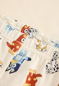 Baumwollshorts mit einem cremefarbenen Hintergrund, der farbenfrohe Cartoon-Hundemuster in Rot, Blau, Schwarz und Gelb zeigt. Knopfdetails am Bund.