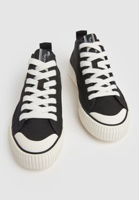 Černé plátěné tenisky s bílými šňůrkami a gumovou podrážkou. Mají kulatou špičku a texturované detaily, s logem na jazyku.