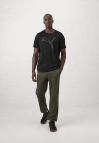 Sort kortærmet t-shirt med et hvidt Puma-logo, parret med olivengrønne bukser og sorte sko med gummisåler.