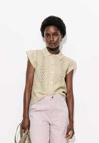 Blouse sans manches en tricot beige avec col et fermeture à boutons, présentant un design ajouré. Associée à un pantalon rayé rose clair.