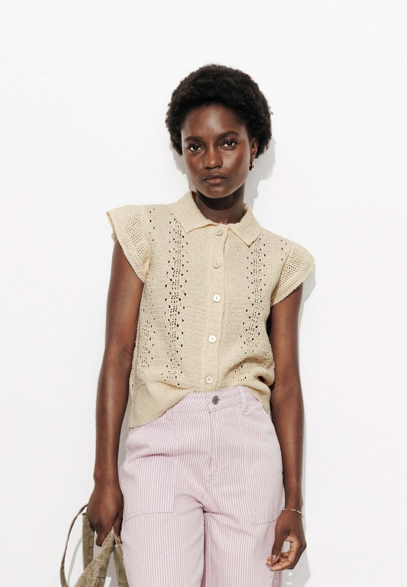 Blouse sans manches en tricot beige avec col et fermeture à boutons, présentant un design ajouré. Associée à un pantalon rayé rose clair.