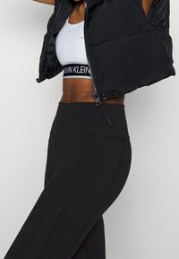 Czarna kamizelka puchowa o pikowanym wzorze, krótka, z zapięciem na zamek, noszona na białym topie z krótkim rękawem z czarną taśmą Calvin Klein oraz legginsami o wysokim stanie.