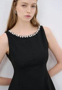 Vestido negro sin mangas con un corsé ajustado y una falda acampanada, con un escote decorativo de cuentas en tonos plateados y transparentes.