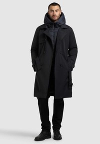 Must särgiga musta trench coat'i, millel on suur kapuuts, kahekordne nööpide rida ja küljetaskud, on kujundatud pehme jakiga. Üksikasjalikud mustad püksid ja saapad.