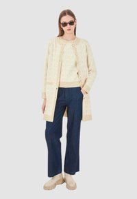 Cardigan a motivi beige chiaro con bottoni, indossato sopra un maglione avorio, abbinato a pantaloni a gamba larga blu scuro e scarpe con zeppa beige.