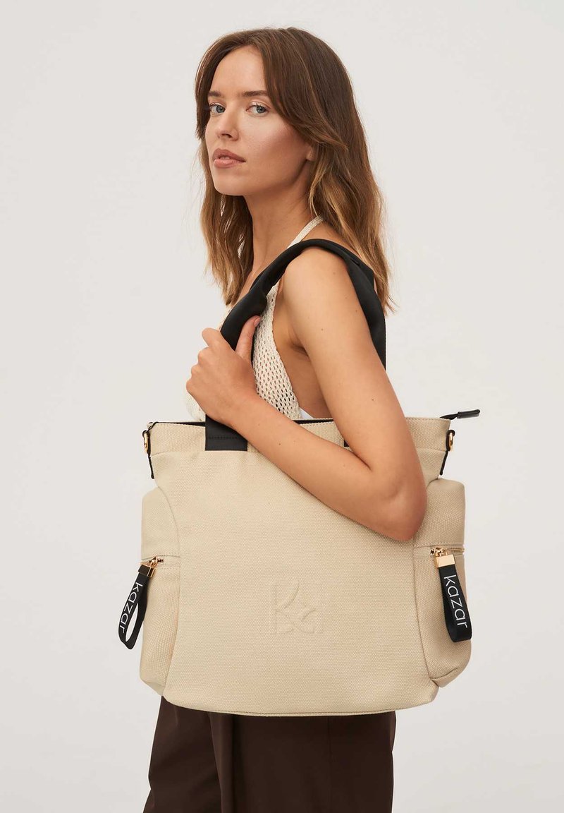 Beżowa torba na ramię z płótna z czarnymi paskami, ozdobiona złotymi zamkami oraz wytłoczonym logo. Zawiera zewnętrzną kieszeń na zamek i teksturowaną powierzchnię.