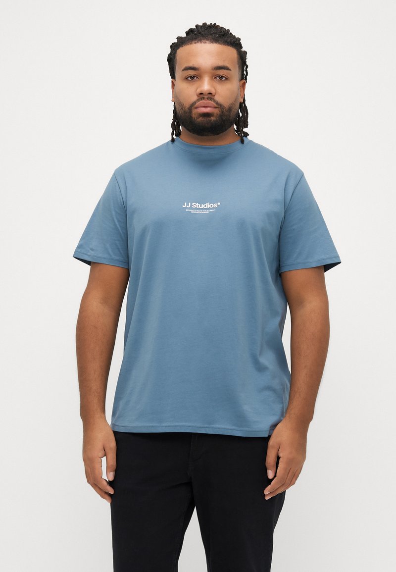 Lyseblå bomulds-T-shirt med rund halsudskæring og korte ærmer; har hvidt "JJ Studios"-logo trykt på forsiden. Enkel design.