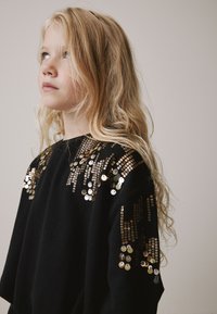 Črni pulover s kapuco z zlatimi sequinami na ramenih v vzorčnem dizajnu. Tkanina je videti mehka, z ohlapnim krojem in dolgimi rokavi.