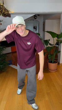 Μακρυμάνικο μπορντό t-shirt με λευκό λογότυπο, γκρι παντελόνι, ασημί αθλητικά παπούτσια και ανοιχτό μπεζ καπέλο. Εσωτερικός χώρος με φυτά και έπιπλα.