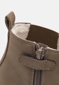 Brązowy zamszowy botek sięgający kostki ma teksturowane wykończenie wnętrza, widoczny zamek błyskawiczny oraz pętlę ułatwiającą zakładanie. Wzdłuż krawędzi znajdują się detale szwów.