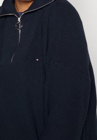 Granatowy sweter dzianinowy z półsuwnym zamkiem i małym logo w kolorze czerwonym, białym i niebieskim na klatce piersiowej w pobliżu ramienia.