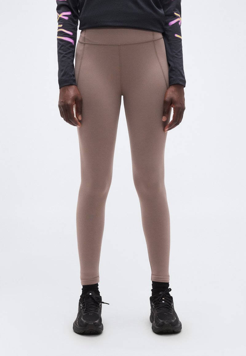 Άτομο που φοράει κολάν σε αποχρώσεις ταουπέ, μαύρα αθλητικά παπούτσια και μαύρο μακρυμάνικο τοπ με μοβ και κίτρινες λεπτομέρειες, που στέκεται μπροστά σε λευκό φόντο.