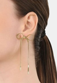 Guldtonade örhängen med en rosett-design och två långa fransar, tillverkade av slätt metall med en stilren yta och subtil textur.